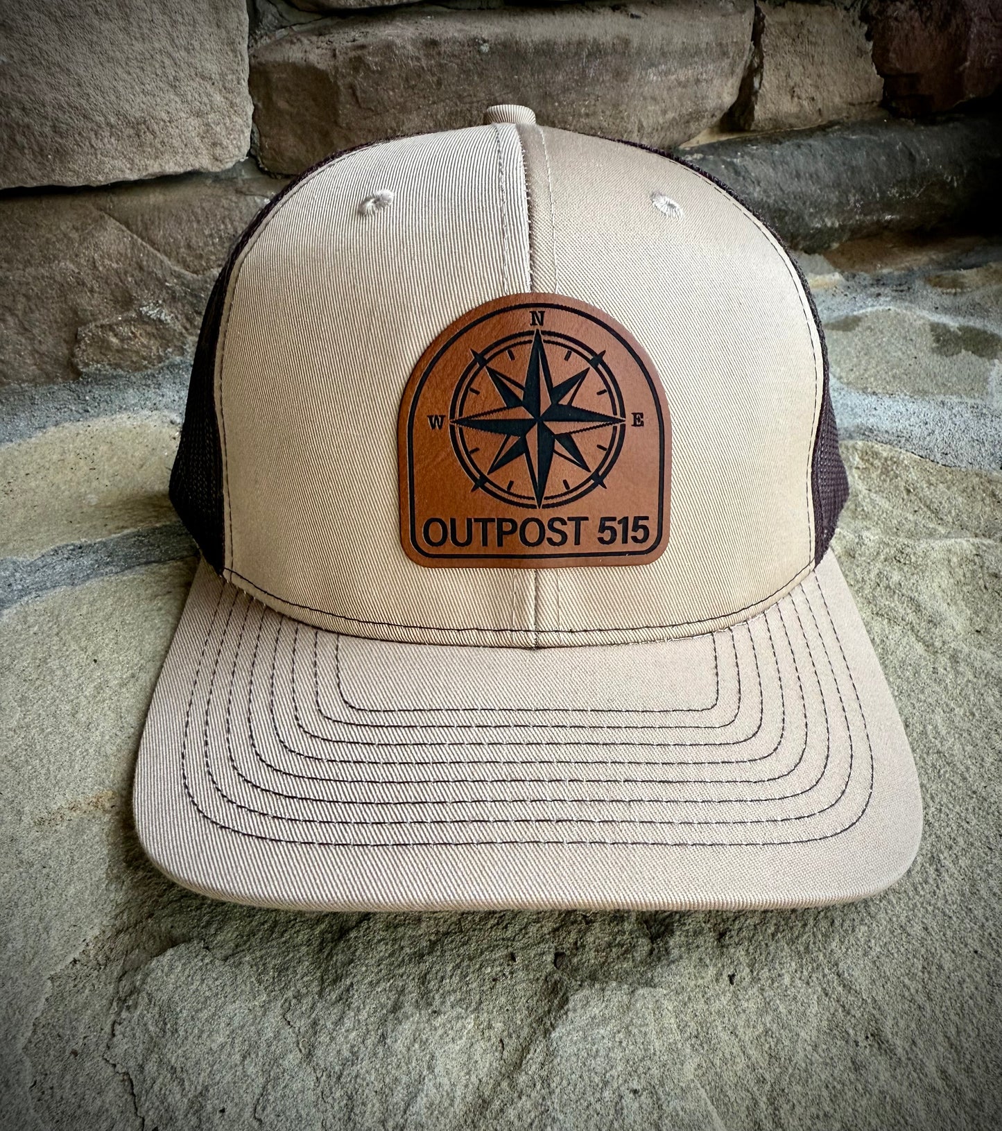 Beige and black cap with a leather patch featuring a compass design and 'Outpost 515' text, placed on stone steps.