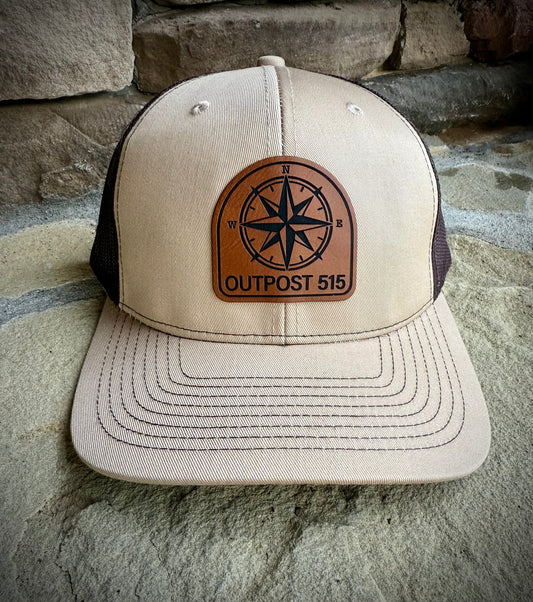 Beige and black cap with a leather patch featuring a compass design and 'Outpost 515' text, placed on stone steps.